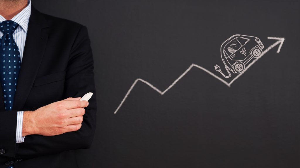 A businessman in a suit holds chalk, standing next to a blackboard with a rising graph drawn on it. An outline of an electric car with a charging plug is drawn ascending the graph, pointing towards an upward arrow, symbolizing growth in the EV market.