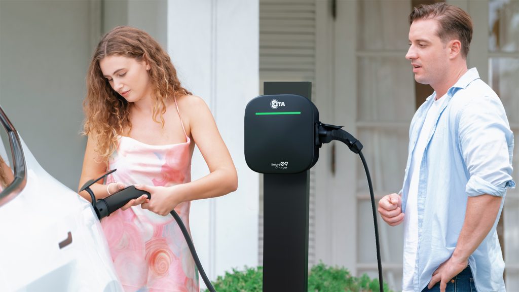 The image shows a woman in a light-colored dress plugging a charging cable into a white electric vehicle, which is partially visible on the left. Next to her, a man in a light blue shirt stands and watches.