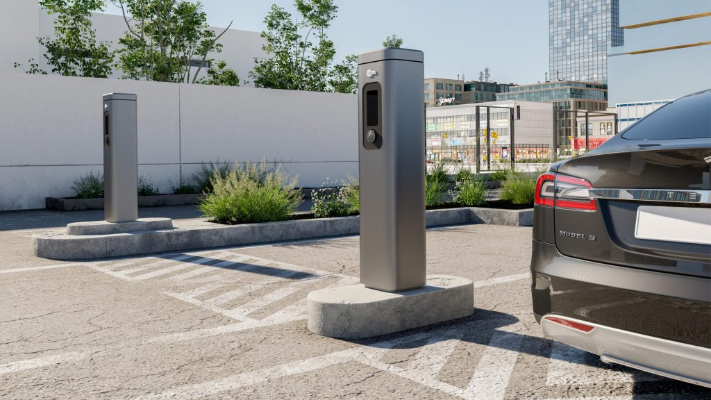 The image displays an outdoor parking lot with two modern, tall gray EV charging stations. One station is in the foreground, and another is further back to the left.