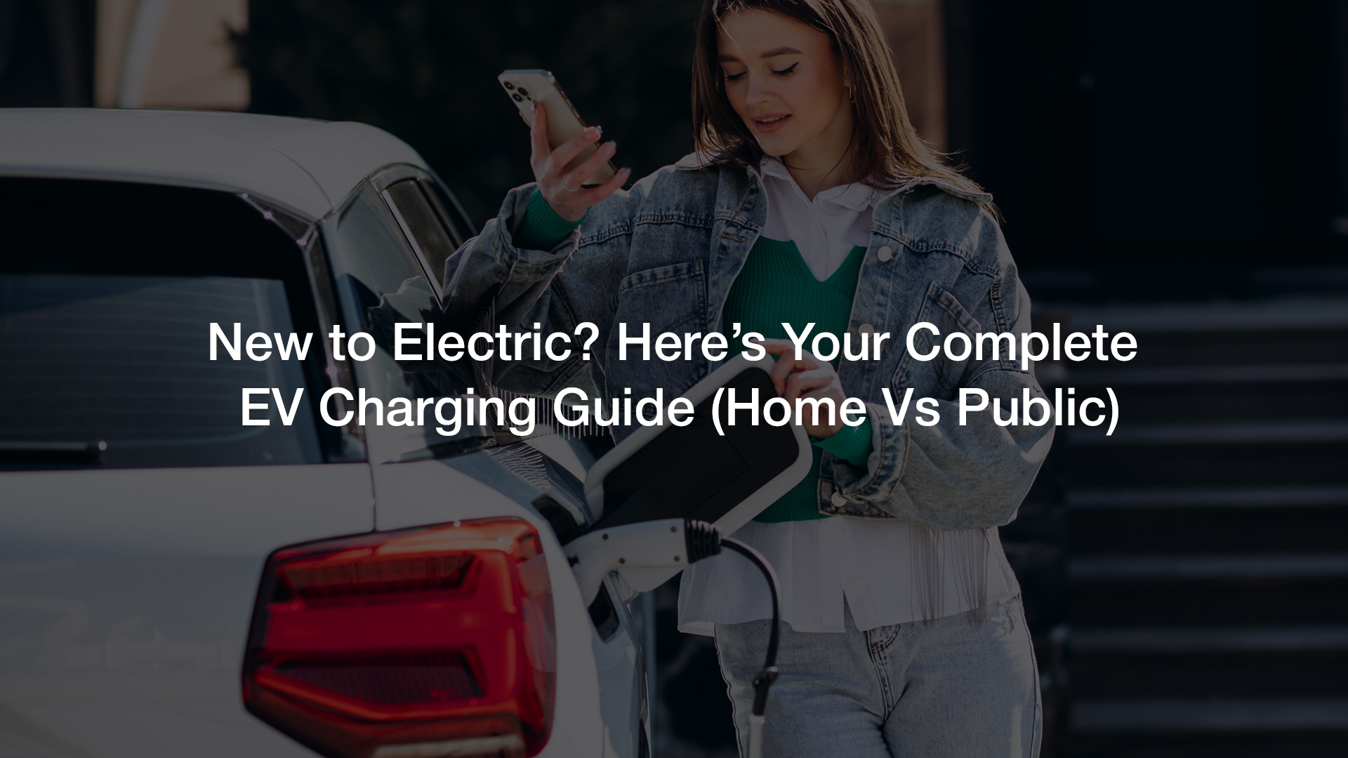 The image shows a person, from the chest down, wearing a denim jacket over a green sweater and light-colored jeans, holding a white EV charging cable and preparing to plug it into the charging port of a white electric vehicle.